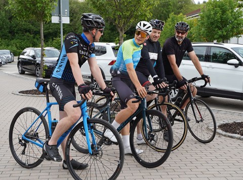 Eine Gruppe mit vier Fahrradfahrern ist zu sehen. Sie unterhalten sich noch und sind kurz davor los zu fahren. Alle tragen Fahrradbekleidung und Fahrradhelme. 