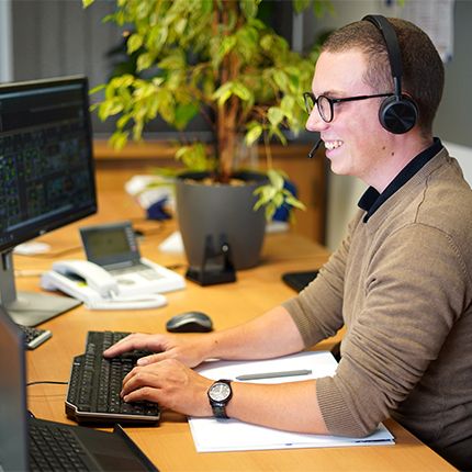 Ein junger Mann mit Brille und braunem Pullover sitzt im Büro vor seinem Rechner. Er trägt ein Headset.
