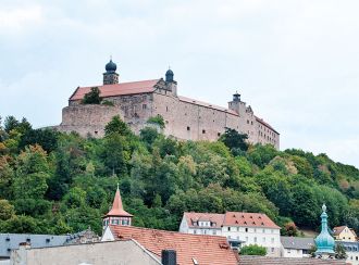 Blick auf die Plassenburg von Kulmbach. Zu sehen ist die Burg, davor viel grüner Wald. Darunter liegt die Stadt Kulmbach. Zu sehen sind Häuserdächer und zwei Türme.