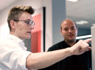 Zwei junge Studenten sind auf dem Bild zu sehen. Der linke Student in weißem Hemd trägt eine Brille und deutet mit dem Finger auf das Display einer Wärmepumpe. Rechts steht ein zweiter Student und schaut aufmerksam zu.