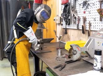 Ein Mann in Schutzmontur (Helm mit Sichtschutz, Schürze, Handschuhe) steht an einem Tisch und hält ein Lötgerät in der Hand. Er ist im Begriff die Arbeit zu starten.