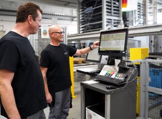 Zwei Produktionsmitarbeiter stehen an einer Blechbearbeitungsmaschine. Sie blicken gemeinsam auf das Bediendisplay. Der linke Mitarbeiter deutet auf das Display und erläutert etwas.