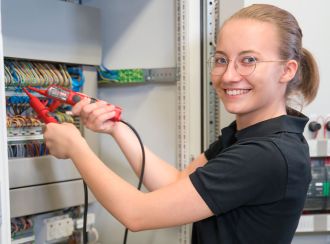 Eine junge blonde Auszubildende von Glen Dimplex Deutschland. Sie trägt eine Brille mit goldener Fassung und ein schwarzes Polo-Shirt.  Die Auszubildende steht mit einem Prüfgerät an einem Wärmepumpen-Schaltkasten und lächelt in die Kamera