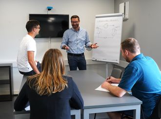 Zu sehen ist ein Schulungsleiter an einem Flip-Chart sowie drei Studenten die vor ihm sitzen und zu hören. Die vier befinden sich in einem Besprechungsraum. Auf dem Flipchart ist der Kältekreis gezeichnet worden.