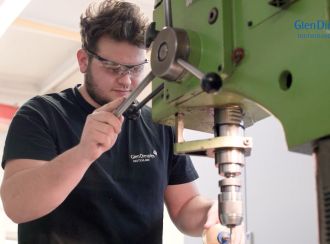 Nick Wildgrube steht an einer Fräsmaschine und arbeitet konzentriert. Er trägt ein schwarzes GDD T-Shirt und eine Schutzbrille.