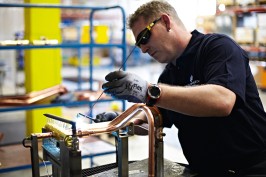 Ein Mitarbeiter aus der Produktion ist bei der Arbeit zu sehen. Er trägt eine schwarze Schutzbrille und hält ein Lötgerät mit dem er kupferfarbene Rohre bearbeitet und verbindet.
