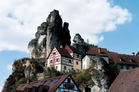 Mehrere Fachwerkgebäude sind zu sehen. Vor einem schmalen hohen Felsen in der fränkischen Schweiz.