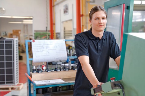 Ein junger Industriemechaniker ist an seinem Arbeitsplatz zu sehen. Er ist umgeben von Maschinen und Werkzeugen.