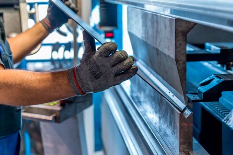 Zu sehen ist eine Werkzeugmaschine. Ein Blech wird gerade gekantet. Ein Mann mit Arbeitshandschuhen hält das Blech, während die Maschine kantet.