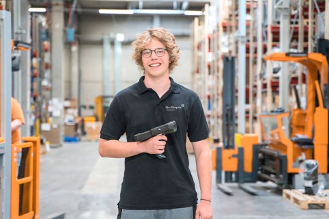 Ein junger Mitarbeiter aus der Logistik steht zwischen Lager-Regalen. Im Hintergrund sind Gabelstapler zu sehen. Er hält einen Scanner in der Hand. 