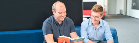 Zwei Studenten sitzen auf einem Sofa und blicken gemeinsam auf ein Buch. 