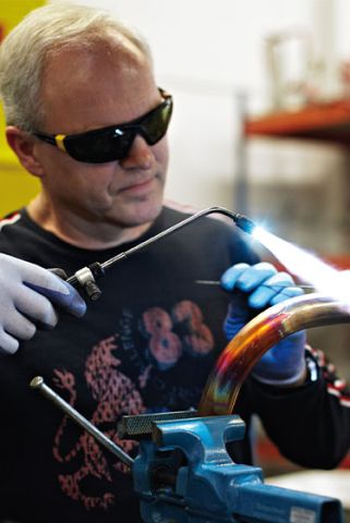 Ein Mitarbeiter aus der Produktion ist bei der Arbeit zu sehen. Er trägt eine schwarze Schutzbrille und hält ein Lötgerät mit dem er kupferfarbene Rohre bearbeitet und verbindet.