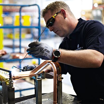 Ein Mitarbeiter aus der Produktion ist bei der Arbeit zu sehen. Er trägt eine schwarze Schutzbrille und hält ein Lötgerät mit dem er kupferfarbene Rohre bearbeitet und verbindet.