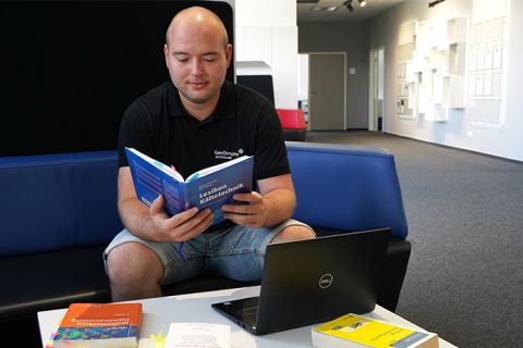 Ein männlicher Student sitzt an einem Tisch und liest in einem Lexikon für Kältetechnik. Vor ihm auf dem Tisch steht ein Laptop und es liegen weitere Fachbücher rum. Er sitzt auf einem blauen Sofa im Forum der Glen Dimplex Deutschland GmbH. 