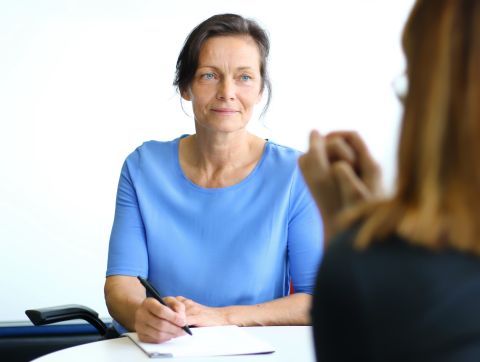 Frau im hellblauen Shirt sitzt am Tisch, hat einen Stift in der Hand, bereit zu schreiben. Hört einer Frau zu, die ihr gegenüber sitzt.