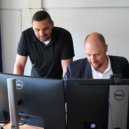Zwei GDD-Mitarbeiter aus der Entwicklung blicken gemeinsam konzentriert in zwei Bildschirme. Einer sitzt, der andere steht neben ihm.