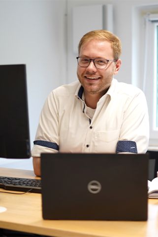 GDD-Mitarbeiter im Büro sitzt am Schreibtisch vor seinem Laptop und Bildschirm und lächelt in die Kamera. Er trägt ein weißes Hemd mit blauem Bund und eine Brille.