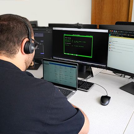 Das Bild zeigt einen Mitarbeiter der IT Abteilung an seinem Arbeitsplatz. Ihm wird über die Schulter geblickt auf einen Arbeitsplatz mit drei großen Bildschirmen und einem weiteren Laptop. Der Mitarbeiter trägt ein Headset.