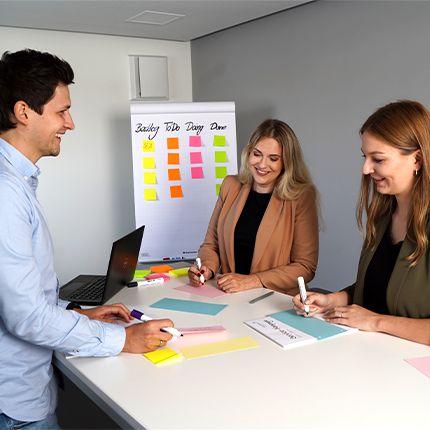 Zwei Mitarbeiterinnen und ein Kollege stehen gemeinsam um einen Tisch, auf dem viele bunte Karten liegen. Sie notieren etwas auf den Karten. Im Hint ergrund steht ein Flipchart, auf dem bereits einige bunte Zettel kleben.