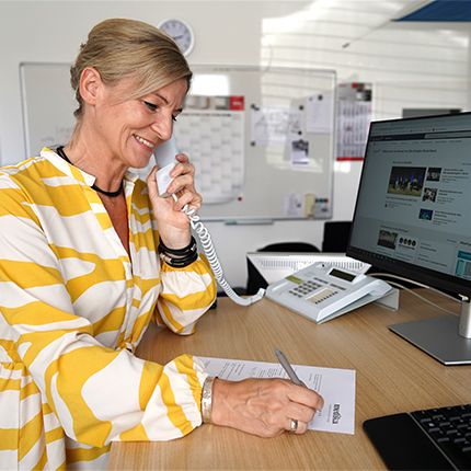 Eine blonde Frau aus der Abteilung Einkauf telefoniert an ihrem Arbeitsplatz. Vor ihr ein Angebot auf dem sie etwas notiert. Sie trägt eine Bluse mit gelb weißem Muster. 