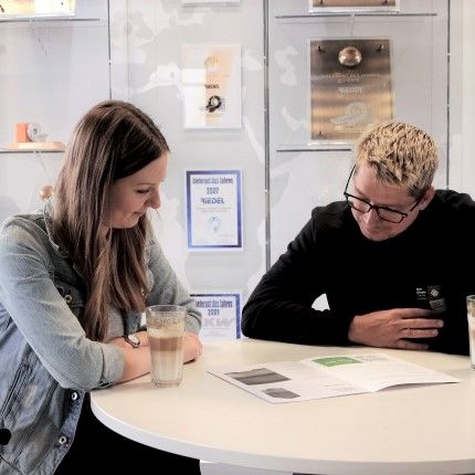 Zwei GDD-Mitarbeiter sitzen am Tisch und blicken in eine Broschüre. Links sitzt eine junge Frau, rechts sitzt ein junger Mann. Vor der Frau steht ein Glas mit Latte Macchiato.